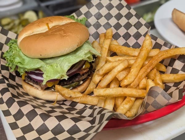 A cheeseburger with lettuce, red onion and pickles beside a basket of fries on checkered paper