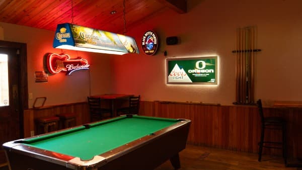 The pool table under a Corona lamp, with Budweiser and Coors Light neon signs on the wall and the cue rack alongside
